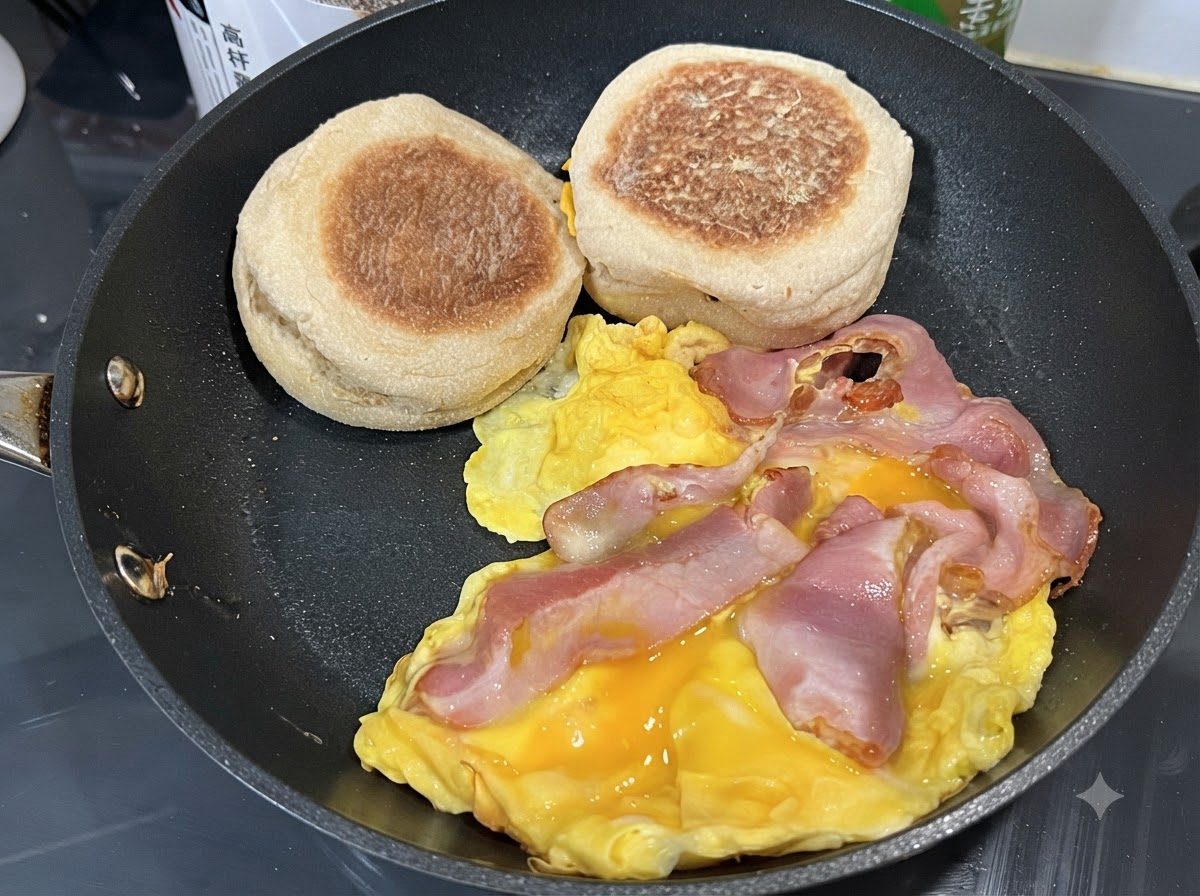 Eggs scrambling in a pan with bacon strips and English muffins resting on the side.