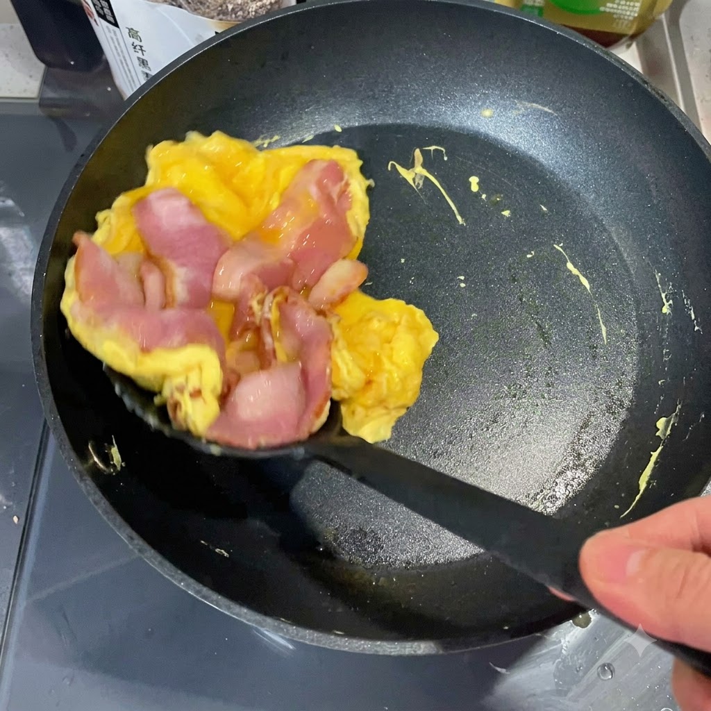 A spatula lifting a cooked egg and bacon stack from a pan to place onto a toasted English muffin half.