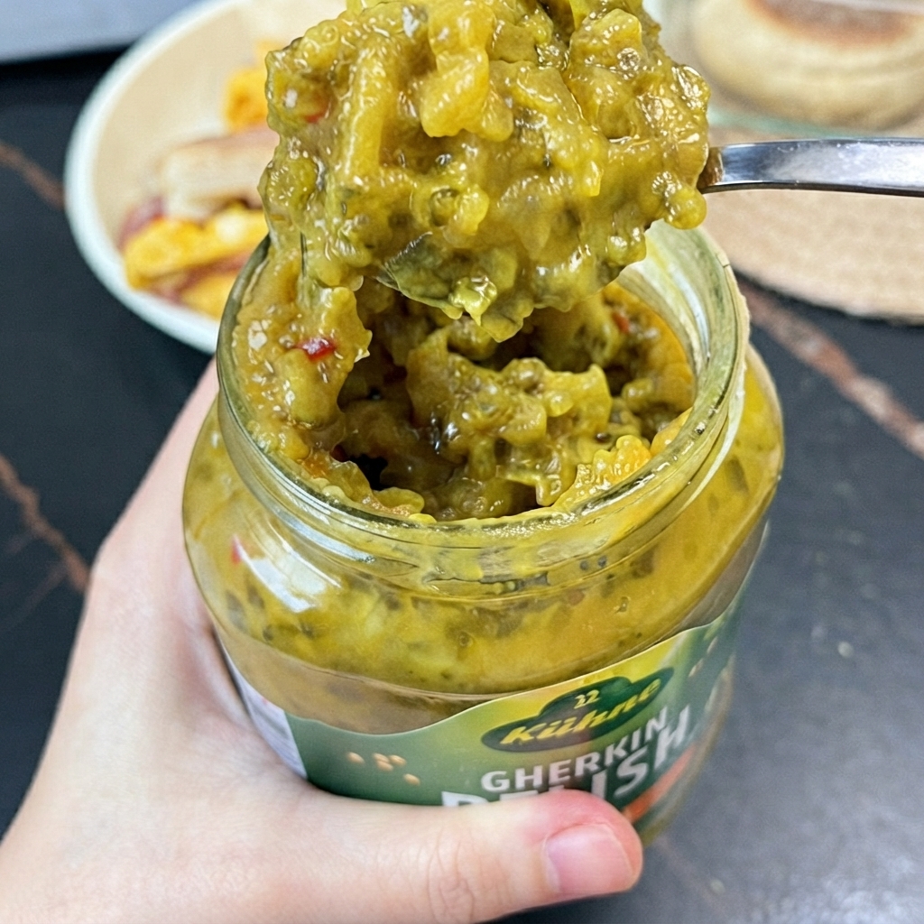 A spoonful of bright green gherkin relish lifted out of a glass jar with a small spoon.