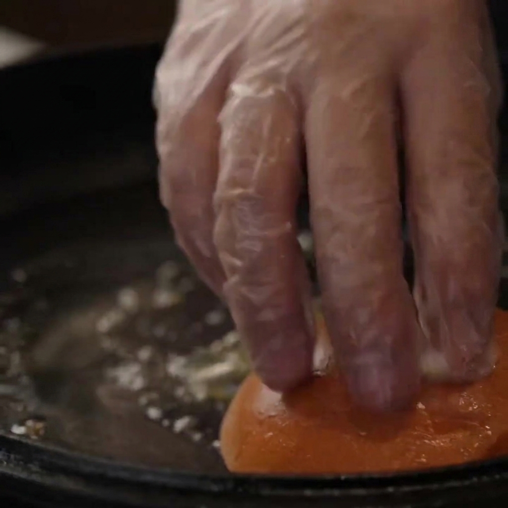 A gloved hand pressing burger buns down into a buttered cast-iron skillet for toasting.