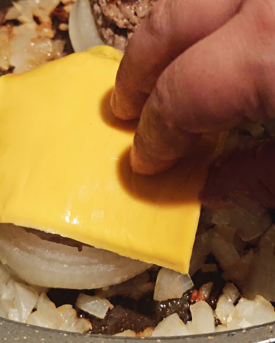 A slice of yellow cheese being placed on top of a cooked meat-stuffed onion ring in the pan.