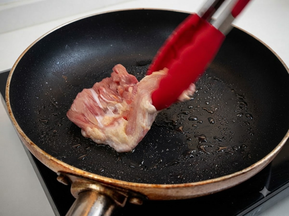 A raw chicken thigh being placed into a heated frying pan with oil using red tongs.