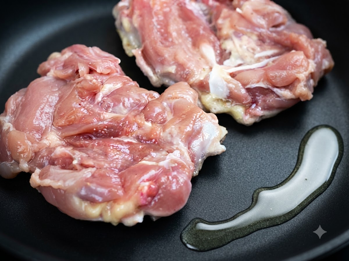 Two raw chicken thighs lying in a frying pan next to a pool of cooking oil.