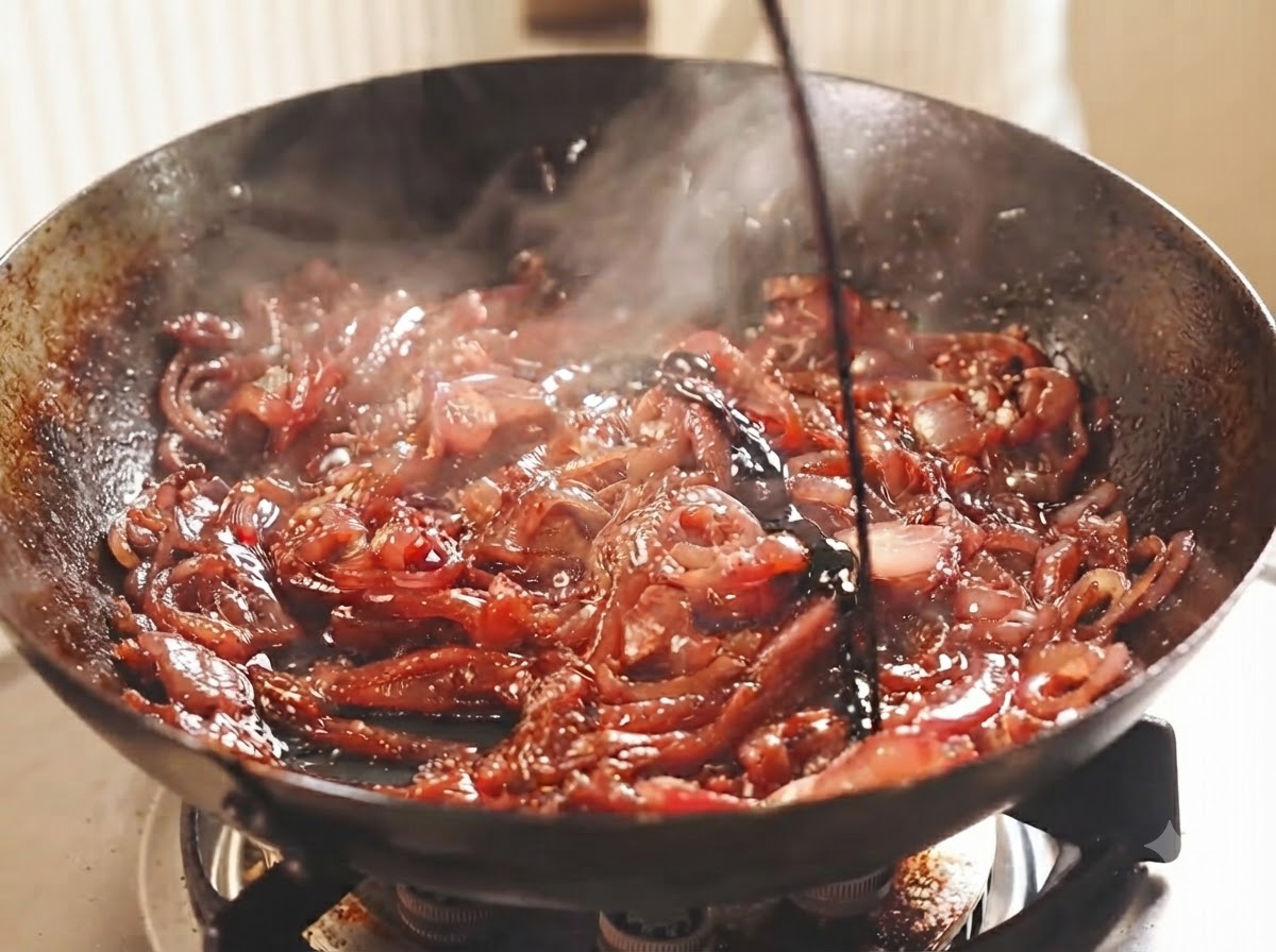 Sliced red onions simmering in a dark wok with dark wine and vinegar being poured over them.