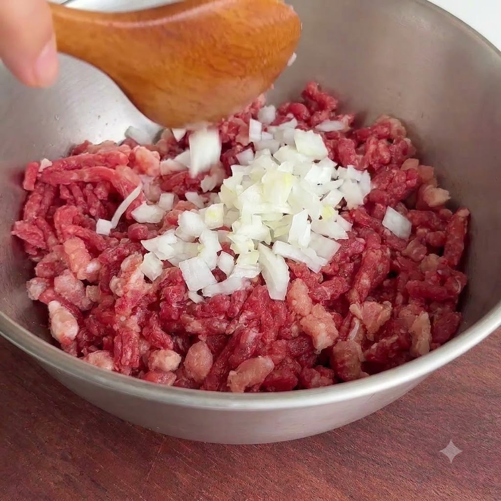 A heap of freshly chopped white onion pieces being dropped from a wooden spoon into a metal bowl of raw minced beef.
