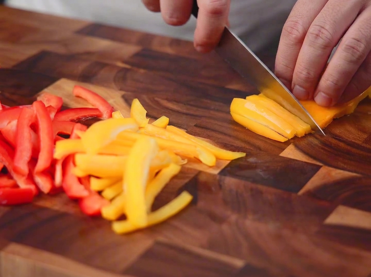 Red and yellow bell peppers being cut into thin strips with a knife on a wooden board.
