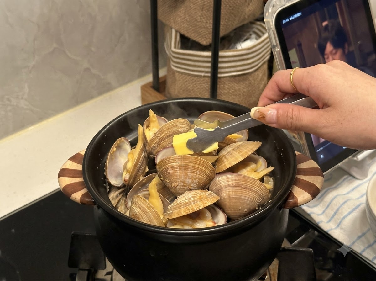 A small piece of butter being placed with tongs into a black pot filled with opened clams.