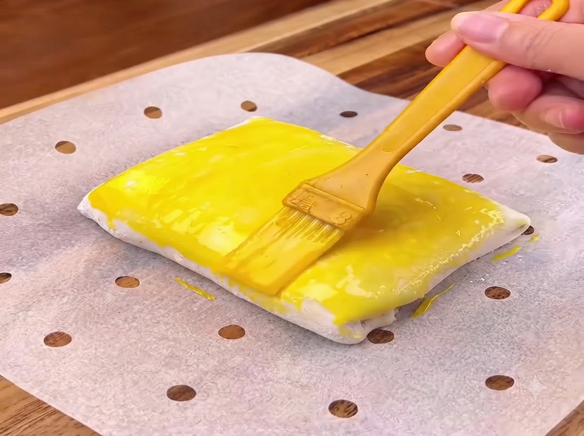 A yellow egg wash being brushed onto the surface of a folded pastry square.