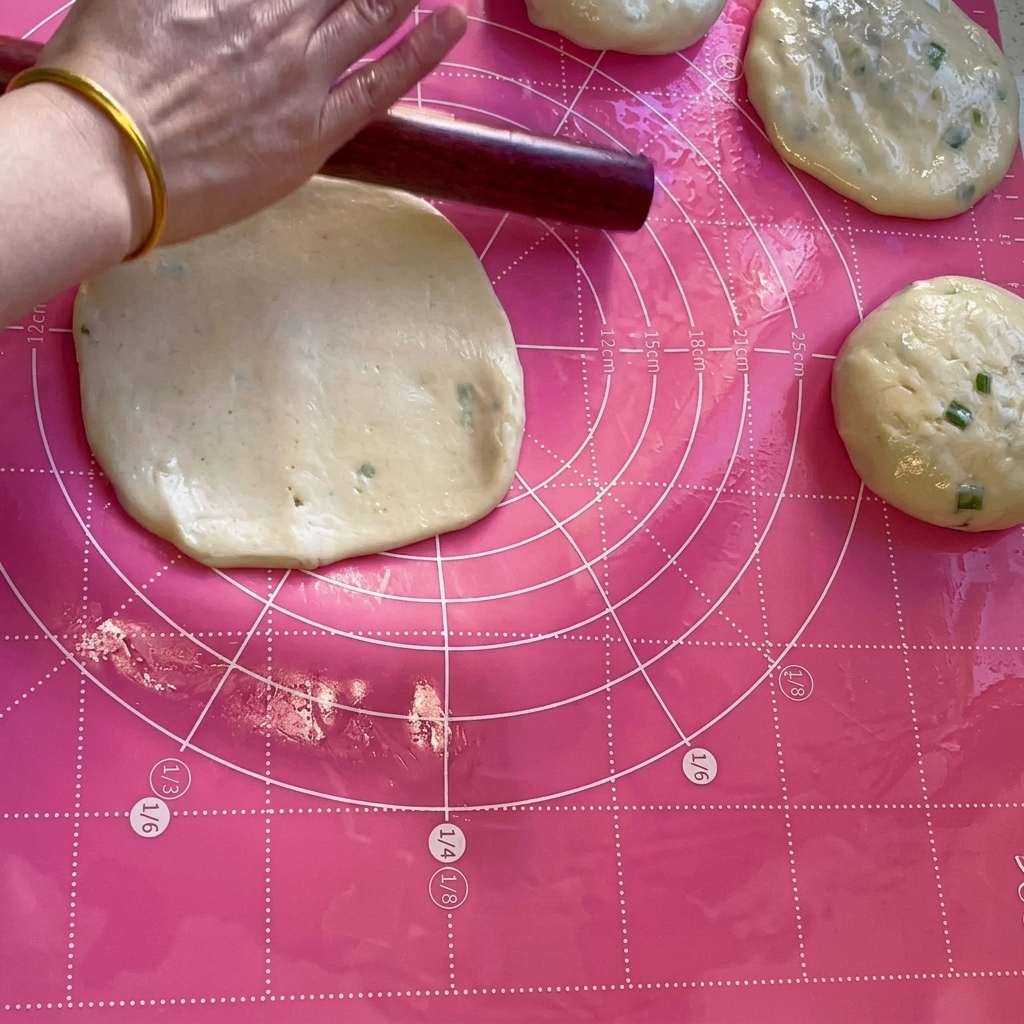 A wooden rolling pin flattening a portion of dough into a thin circle on a pink mat.