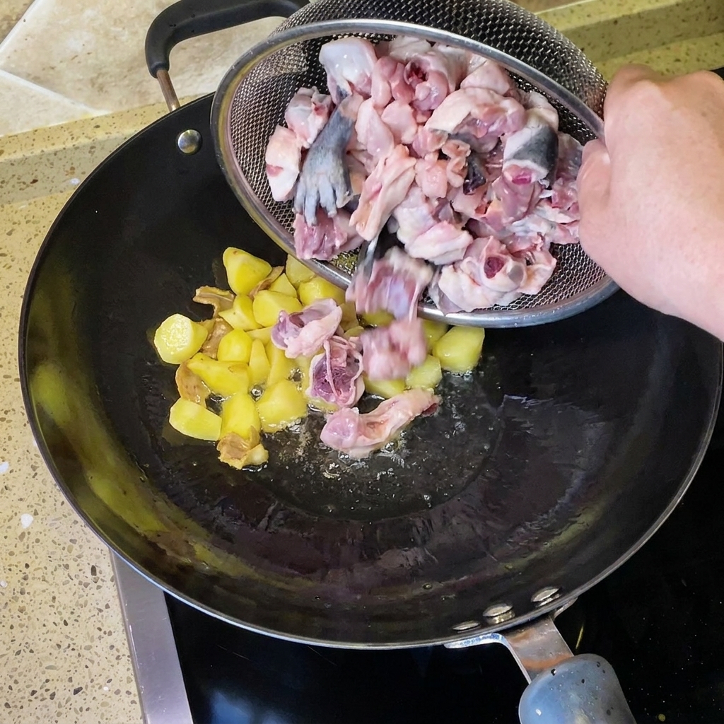 Chopped raw chicken pieces being poured from a wire strainer into the wok with fried potatoes and ginger.