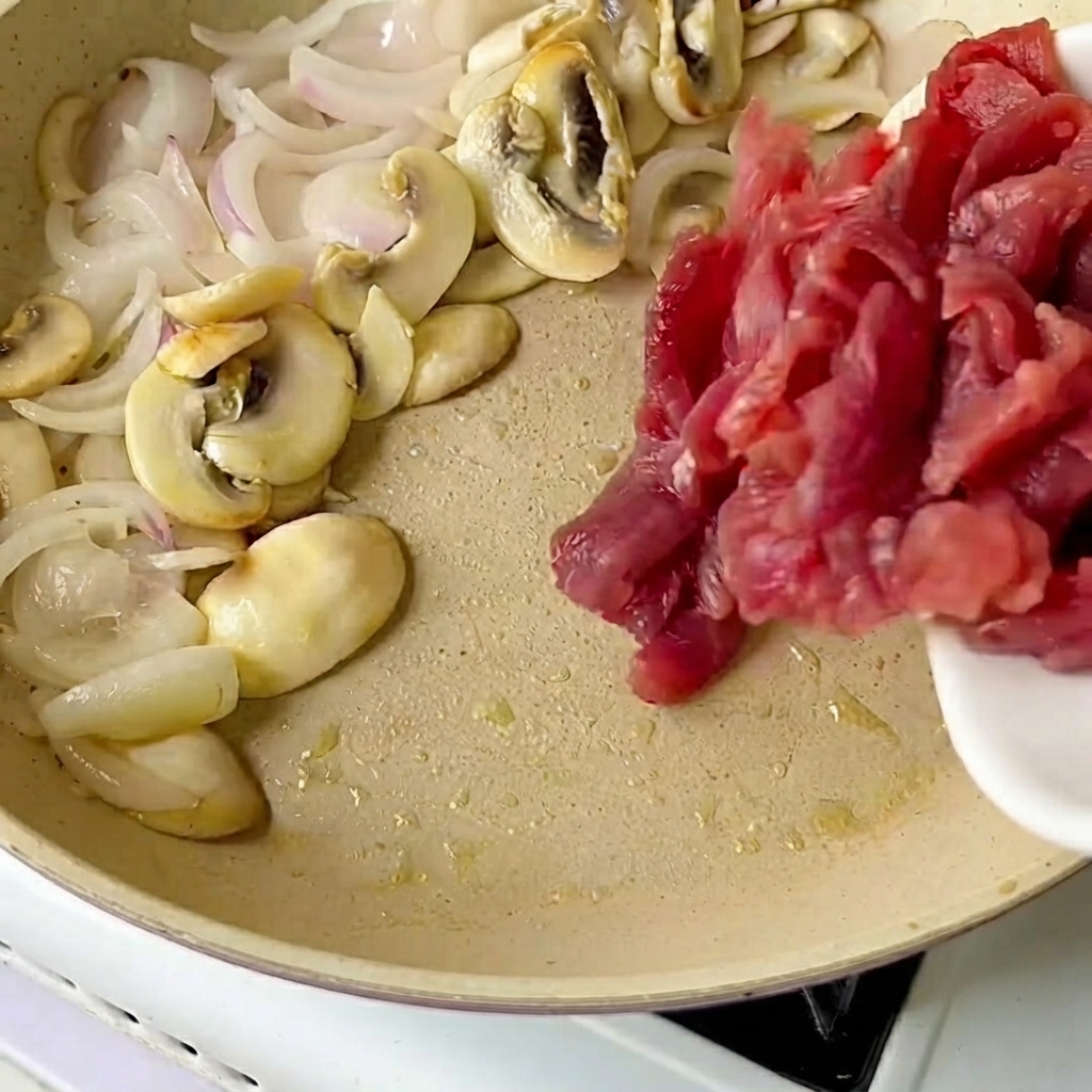 Sliced beef being poured from a white plate into a frying pan containing sautéed onions and mushrooms.