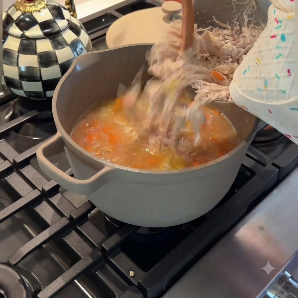 Pouring finely shredded chicken from a metal bowl back into a grey pot of simmering chicken and vegetable soup.