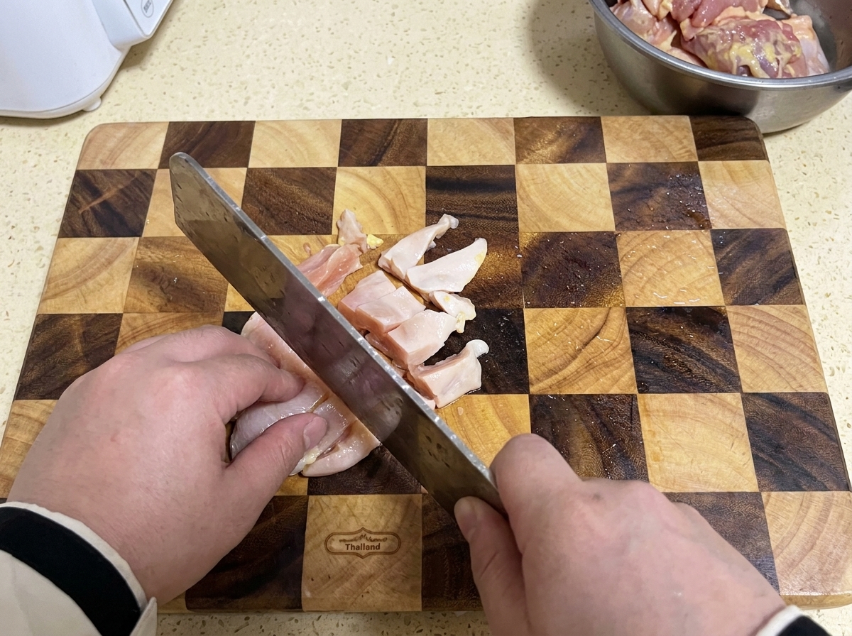 A cleaver slicing raw chicken breast into small pieces on a checkered wooden cutting board.
