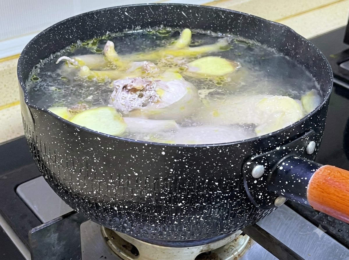 Raw chicken pieces and ginger slices simmering in a speckled black pot filled with boiling water.