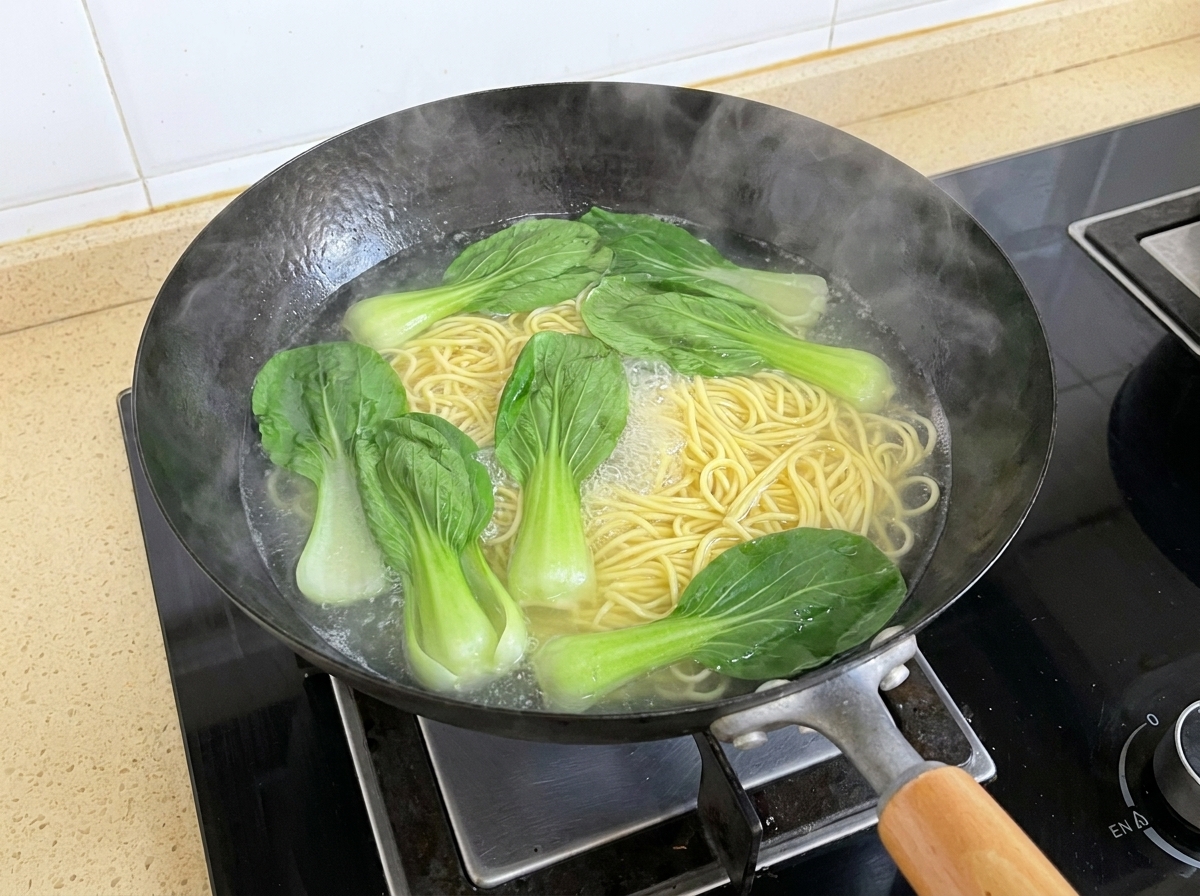 Fresh bok choy leaves floating in boiling water alongside boiling noodles in a wok.