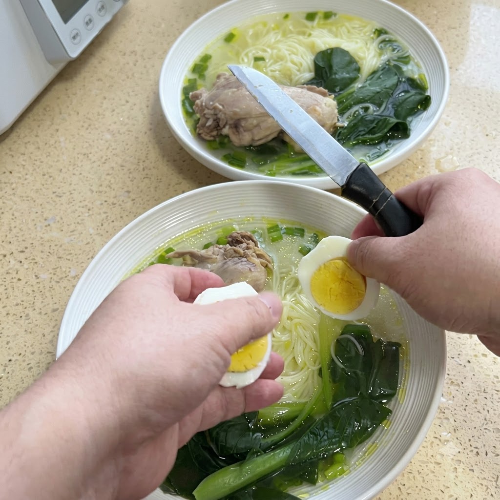 Hands slicing a hard-boiled egg in half with a knife above a bowl of noodles.