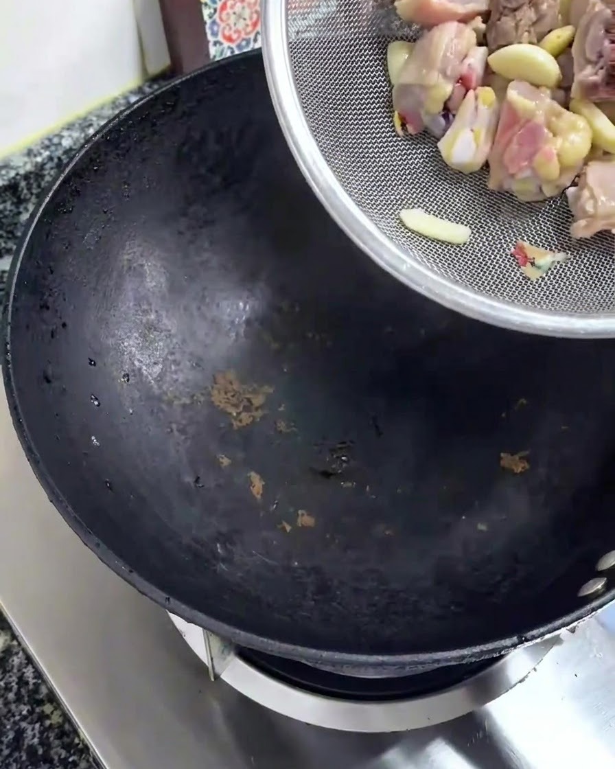 Raw chopped chicken pieces being poured from a silver strainer into a black wok.