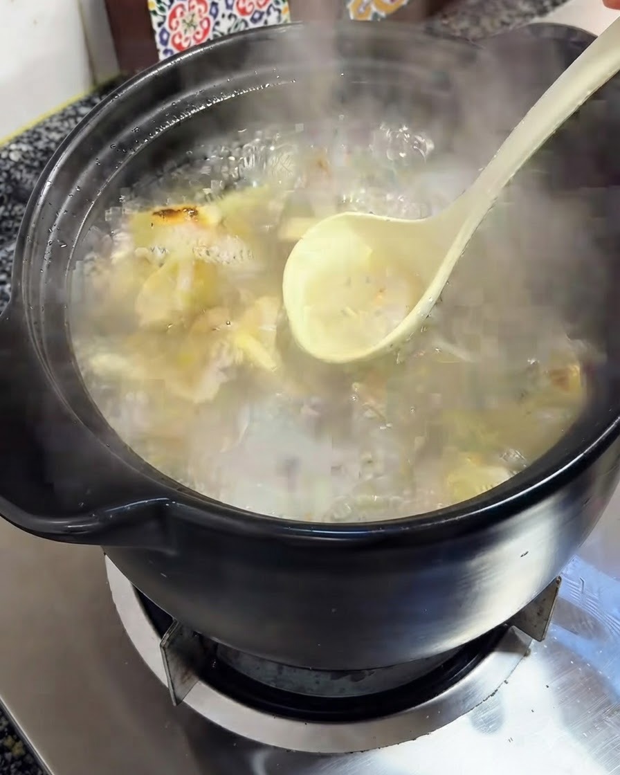 A ladle removing foam from the bubbling broth inside a black clay pot.
