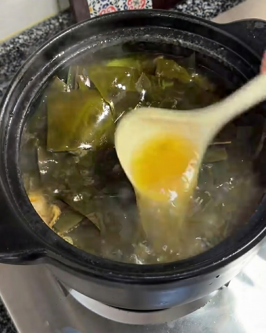 A ladle removing a layer of yellowish oil from the bubbling surface of the soup.