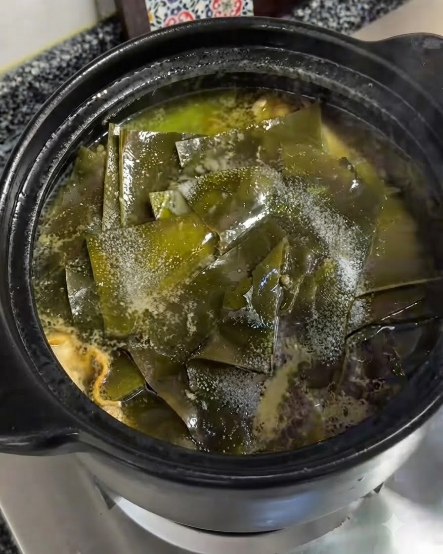 Fine salt being sprinkled directly over the green kelp pieces floating in the simmering clay pot.