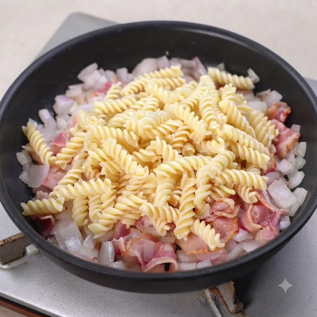Cooked spiral pasta resting on top of cooked bacon and onions in a black skillet.