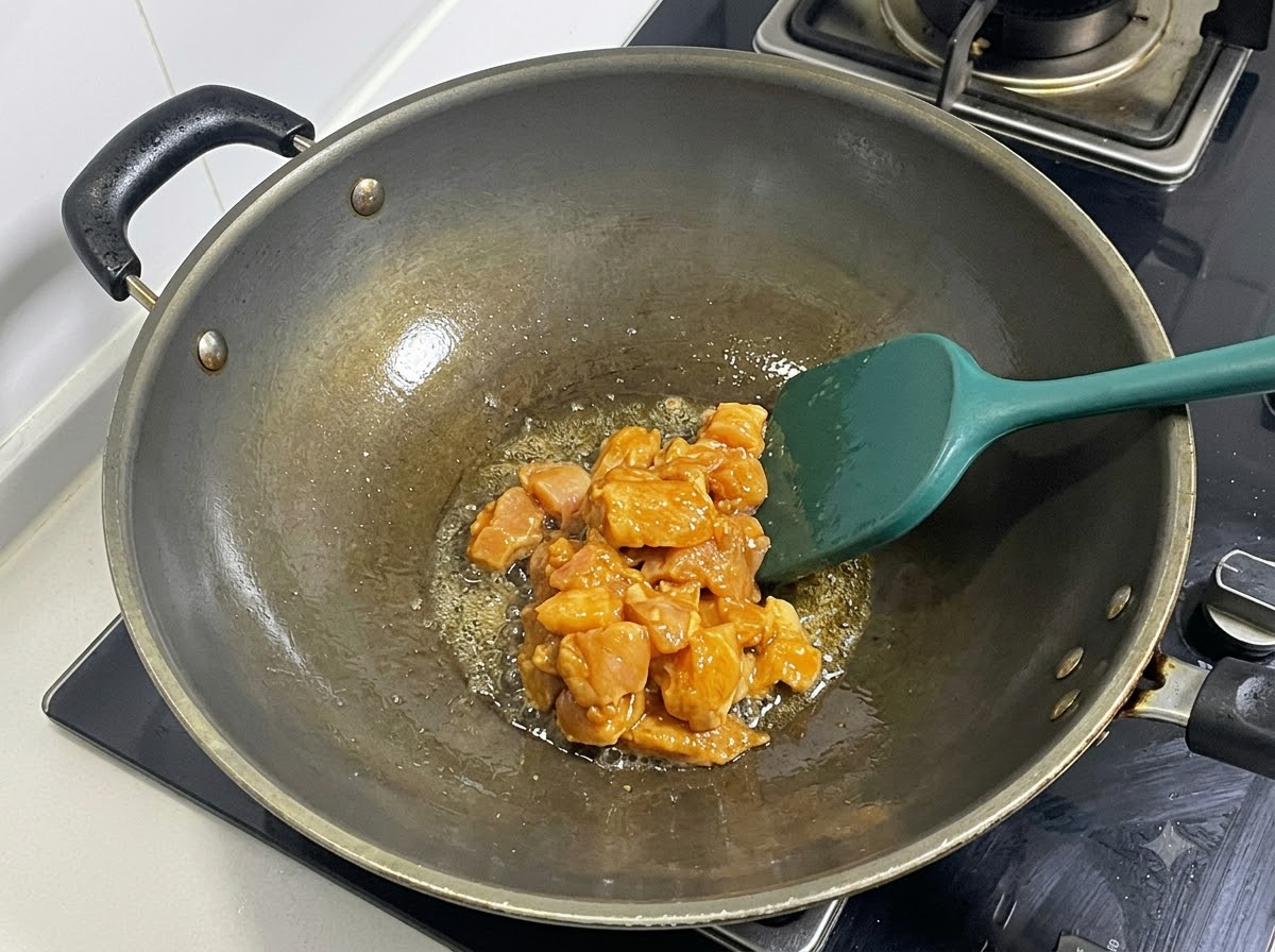 Diced marinated chicken breast being stir-fried in a heated wok with a green spatula.