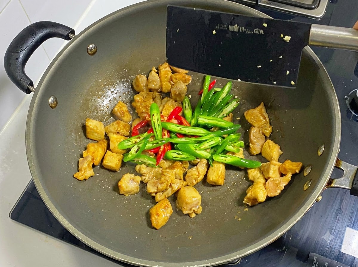 Sliced green and red chilies added to cooked chicken pieces in a wok being stirred with a black spatula.