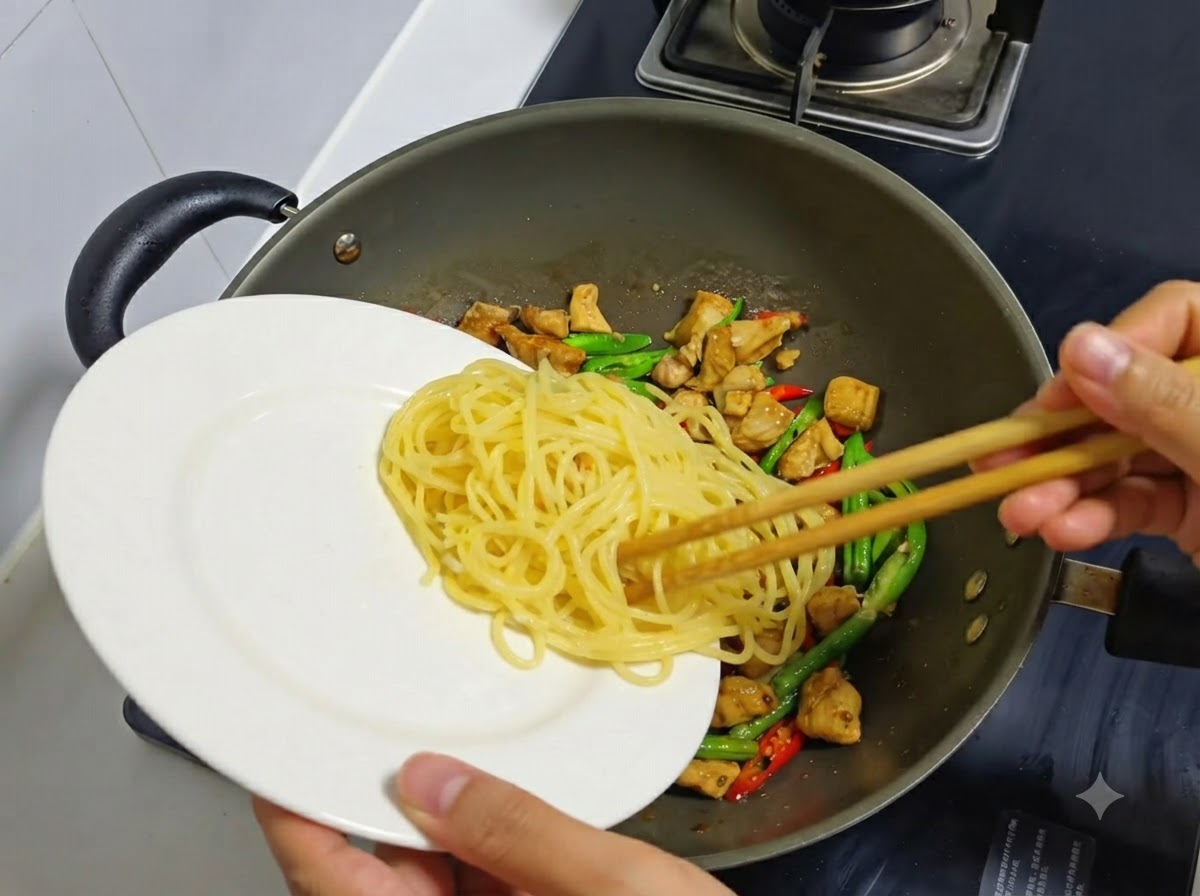 Cooked spaghetti noodles being transferred from a white plate into a wok containing cooked chicken and chilies.