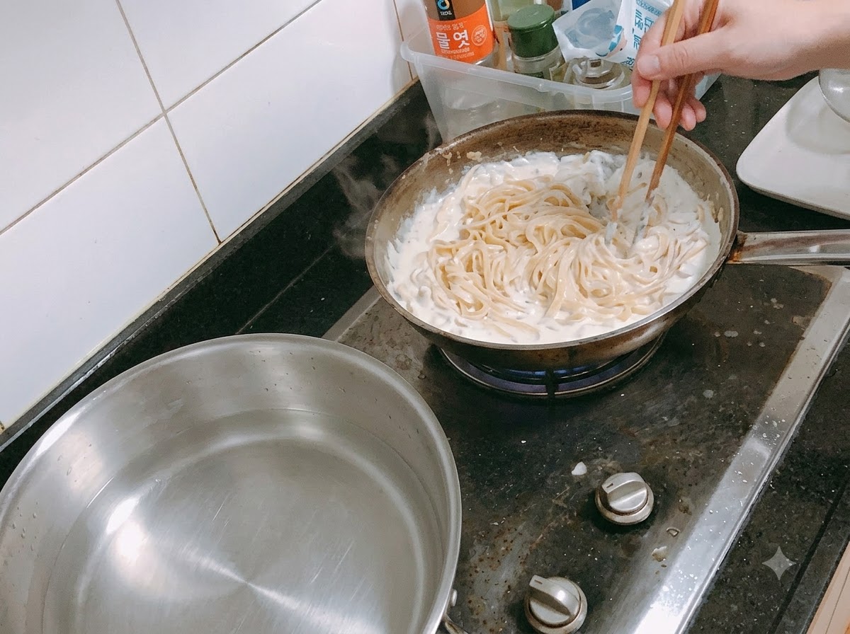 Pasta being mixed into a simmering cream sauce in a pan using chopsticks.