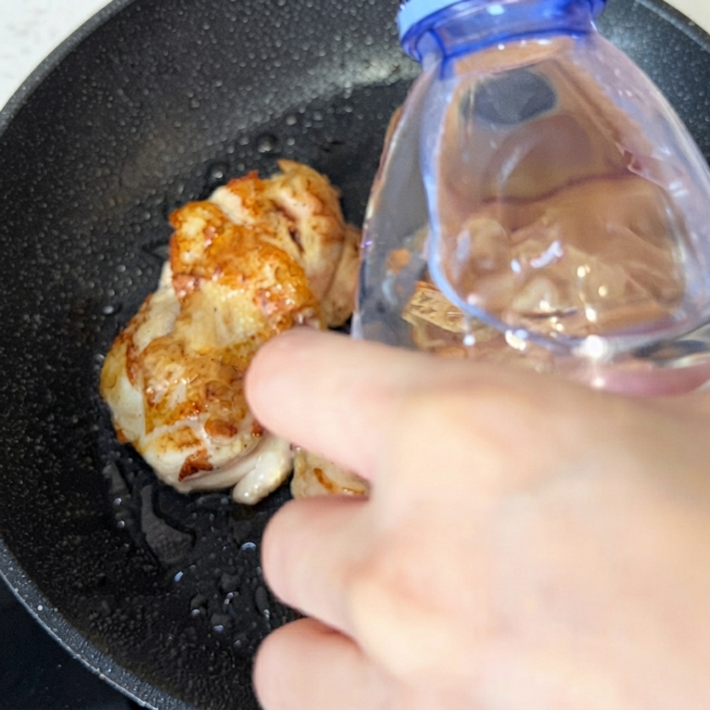 A water bottle pouring a small amount of water into the skillet over cooked chicken pieces.