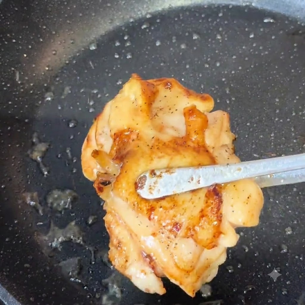 Tongs lifting a golden-brown cooked chicken thigh out of a frying pan.