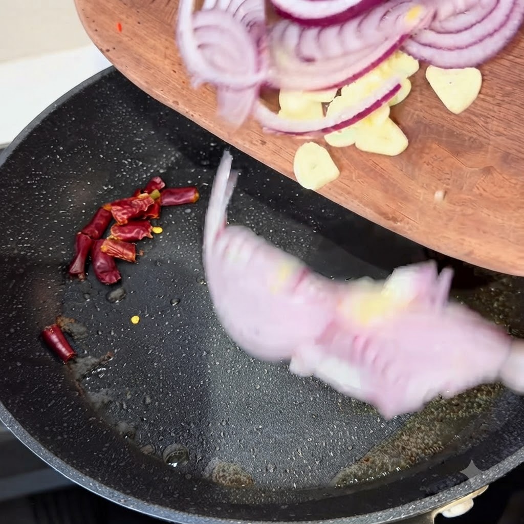 Red onion slices, garlic cloves, and dried chili peppers being transferred from a wooden board into a hot frying pan.