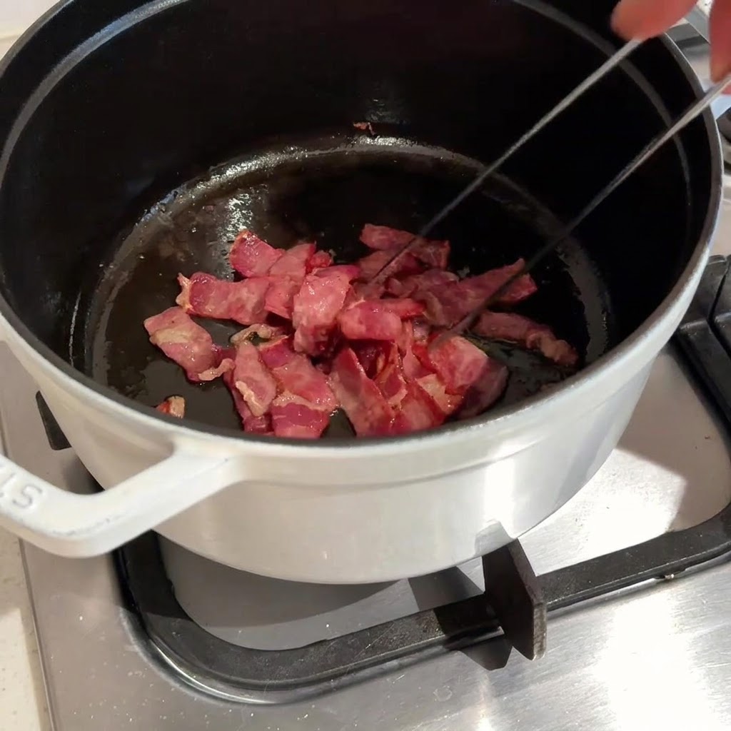 Chopped bacon being tossed and fried in a dark pot using metal tongs.