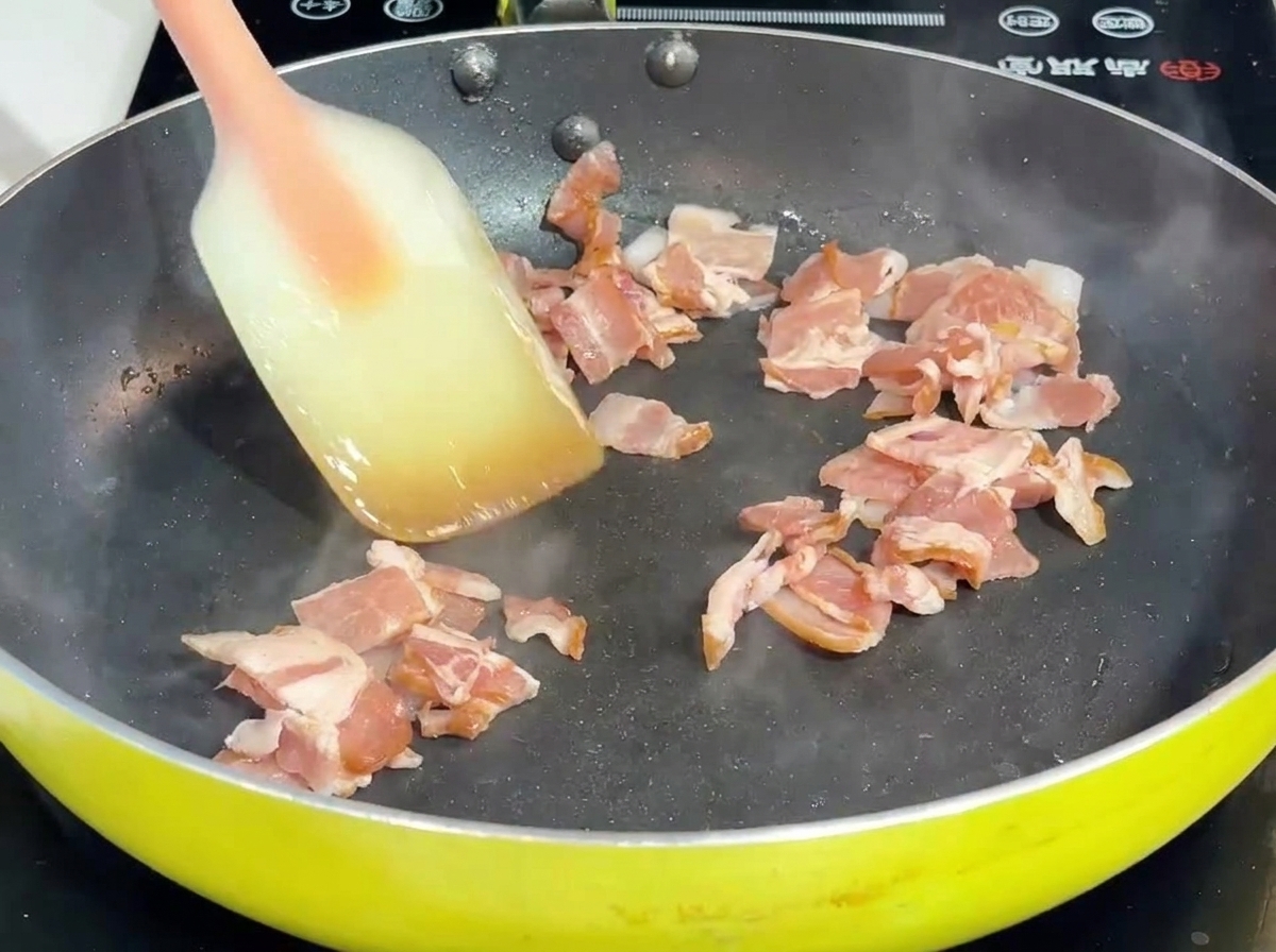Bacon and onion being stir-fried with a spatula in a pan.