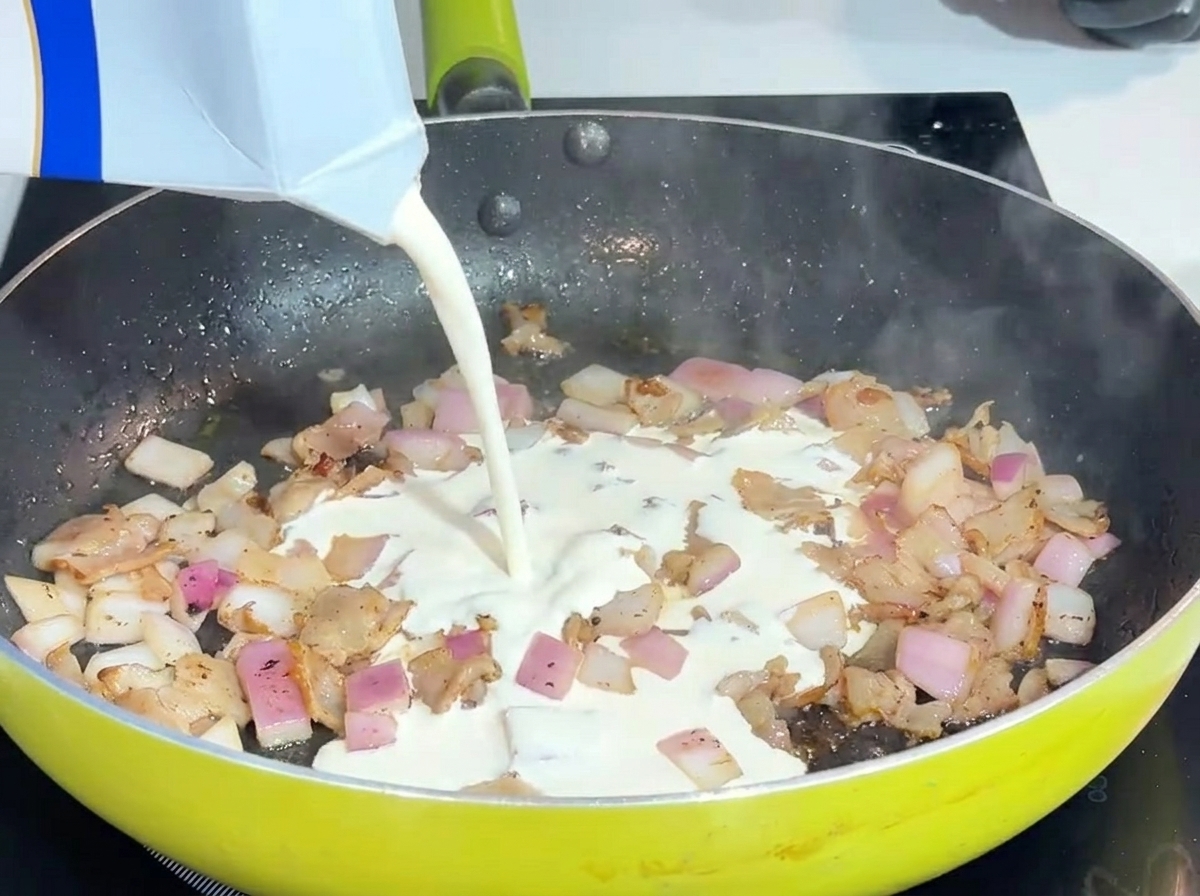 Liquid heavy cream being poured into a pan with bacon and onions.
