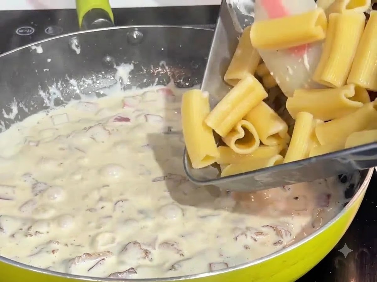 Pre-cooked heart-shaped pasta being poured from a tray into a bubbling cream and bacon sauce in a pan.
