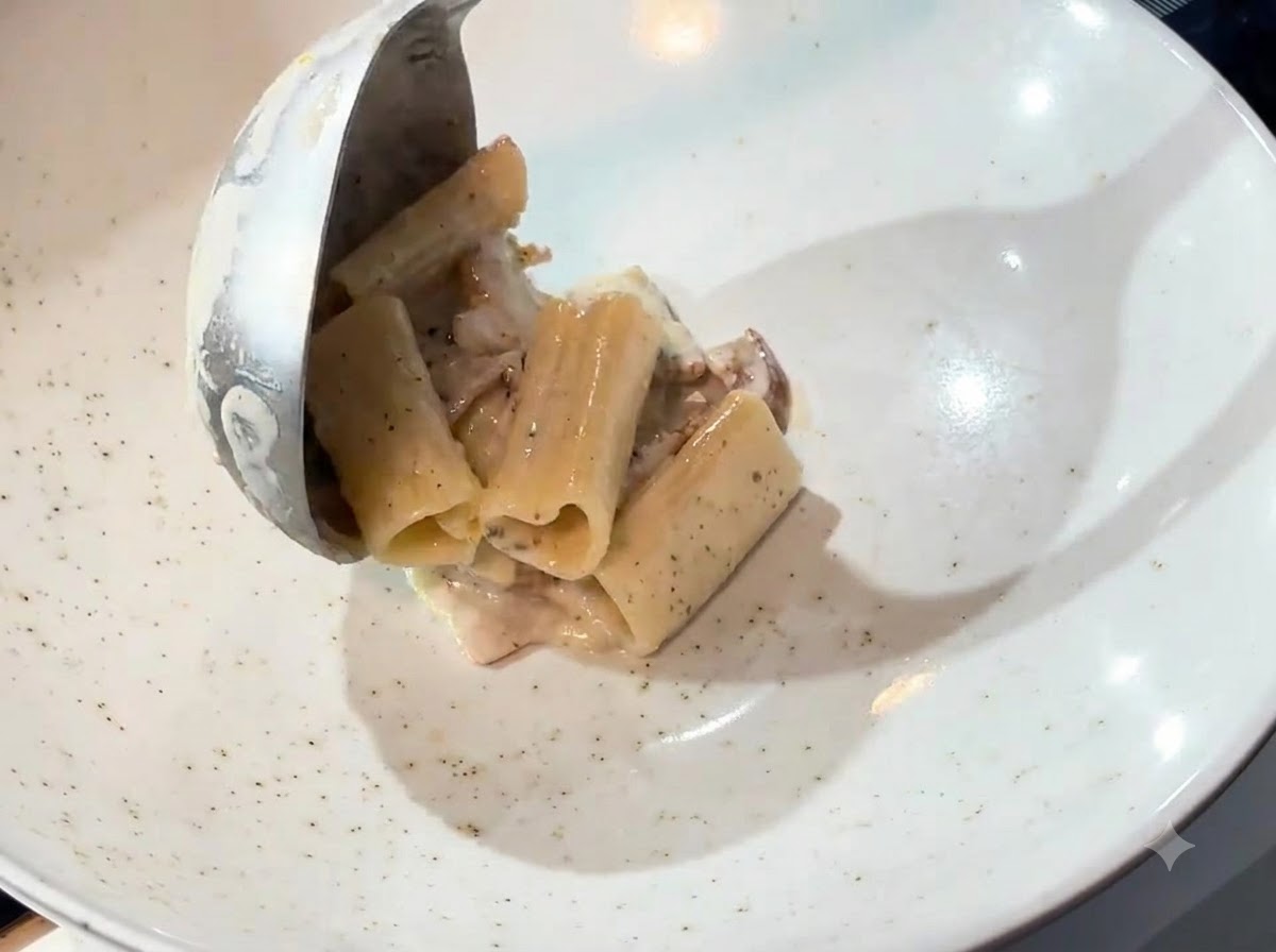 A ladle transferring creamy pasta from a pan into a white serving bowl.