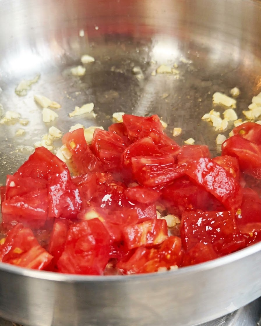 Fresh diced red tomatoes being sautéed with garlic inside a metal pan.