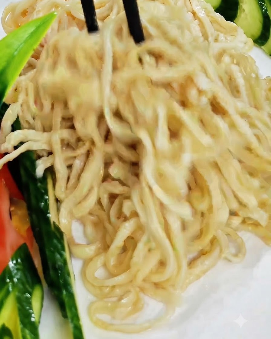 Black chopsticks lifting and arranging cooked noodles onto a white plate garnished with cucumber.