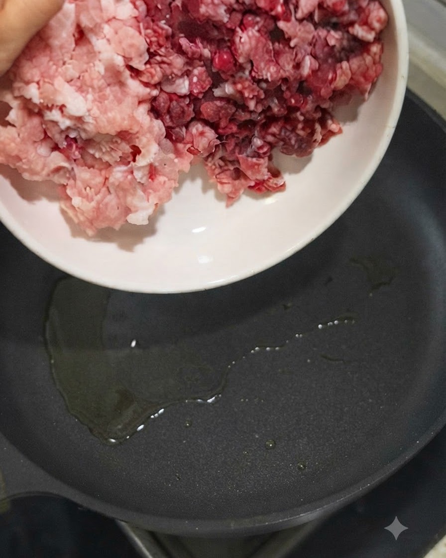 Ground pork and ground beef being poured from a white bowl into a hot oiled skillet.