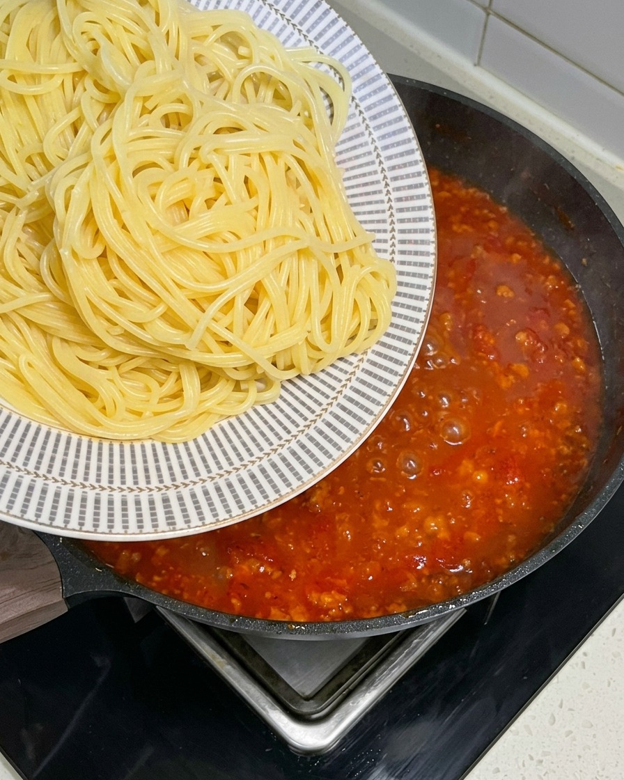 Freshly boiled spaghetti noodles being transferred from a patterned plate into a large pan filled with a bubbling red meat sauce.