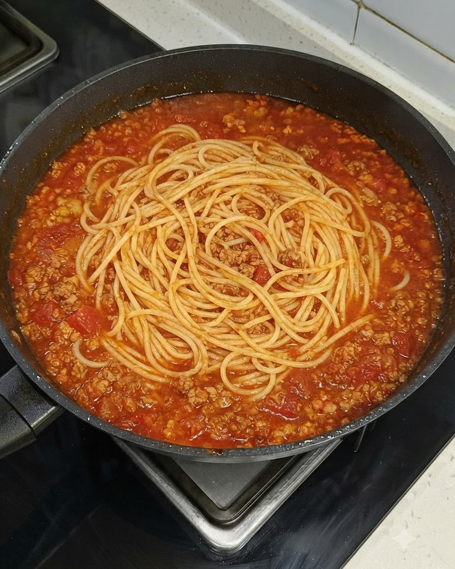 A nest of spaghetti noodles resting evenly inside a large skillet filled with red meat sauce on a stovetop.