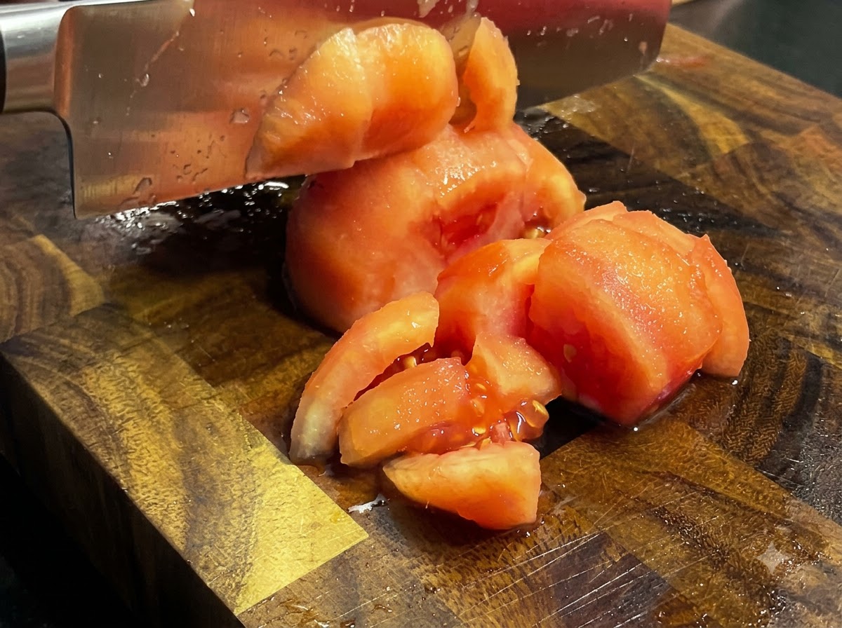 A kitchen knife chopping cooked, peeled tomatoes into chunks on a wooden cutting board.