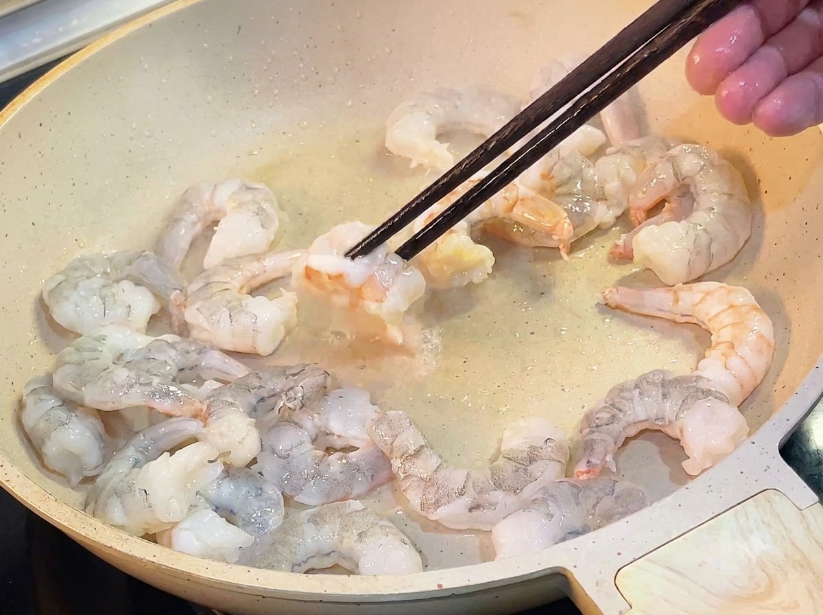Wooden chopsticks flipping pink, cooked shrimp in a lightly oiled frying pan.