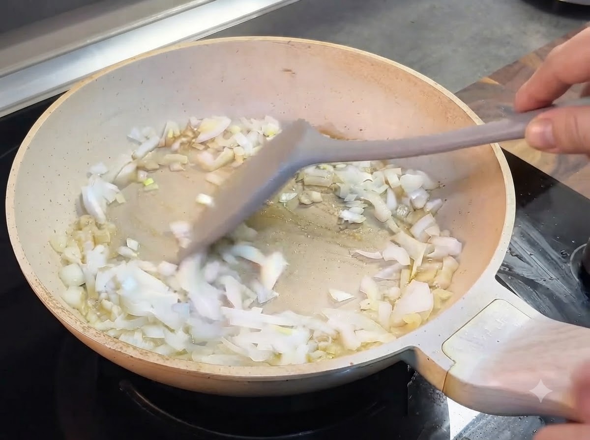 Chopped onions being stirred with a grey spatula inside a light-colored pan.