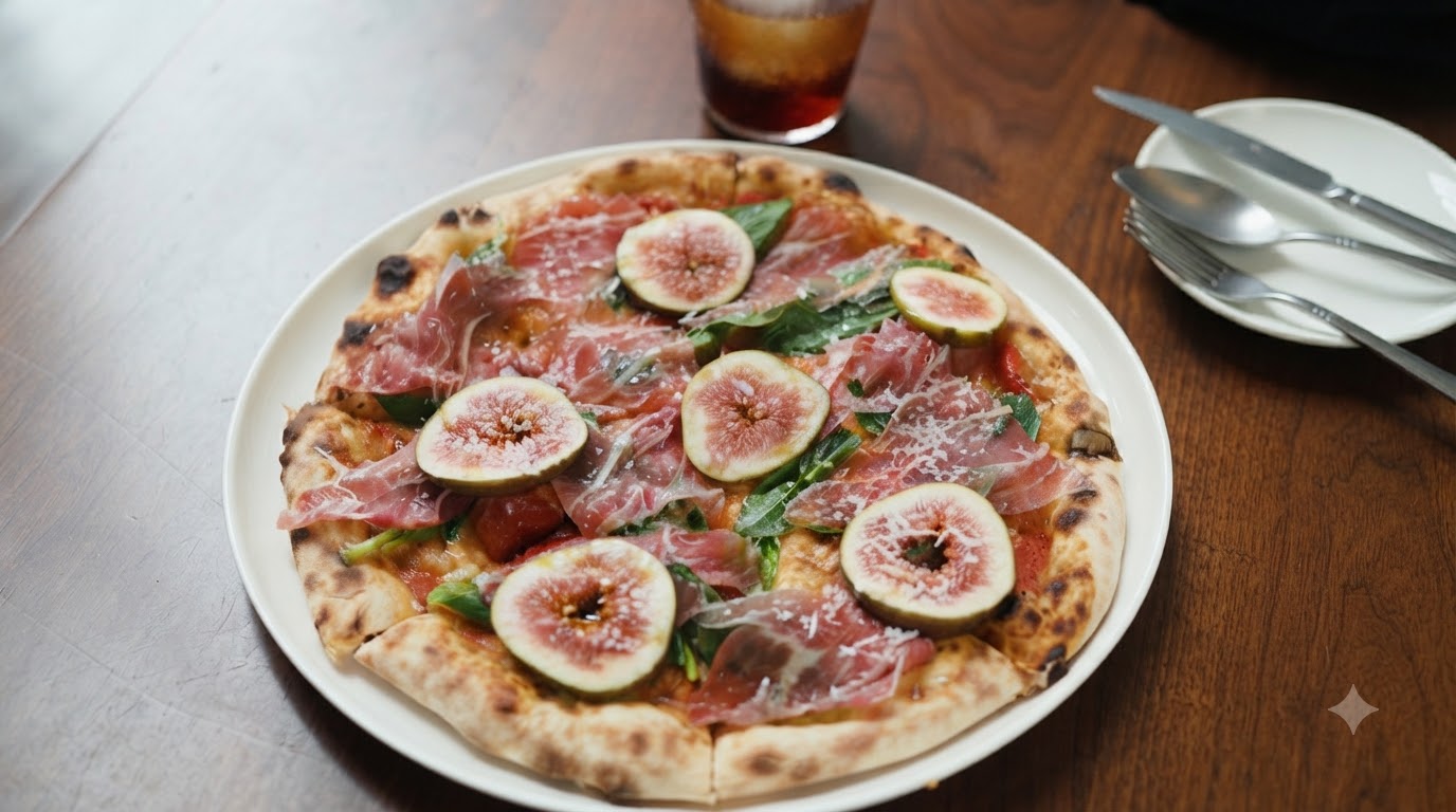 A freshly baked gourmet thin-crust pizza topped with sliced figs, prosciutto, arugula, and grated cheese, served on a white ceramic plate alongside a chilled glass of iced beverage.