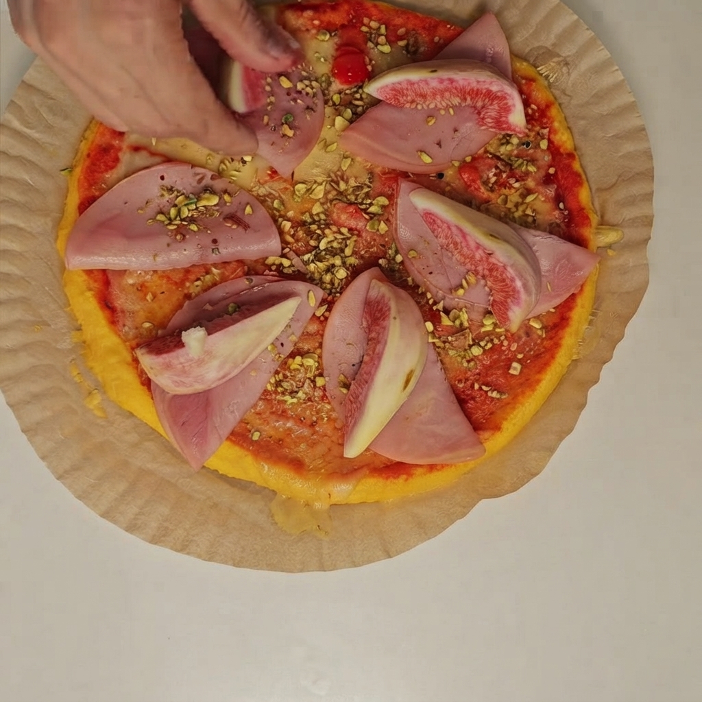 A person placing fresh fig wedges and crushed pistachios over ham slices on the pizza.