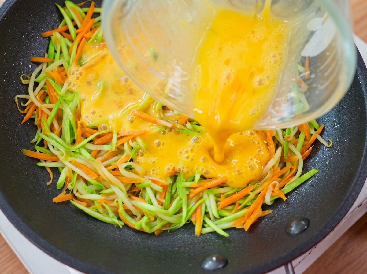 Yellow egg liquid being poured from a measuring cup over the vegetable layer in the pan.