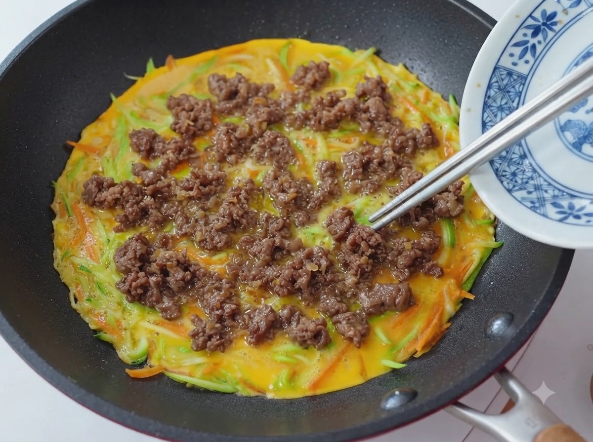 Close-up of chopsticks placing and spreading cooked beef crumbles over a pan with a vegetable and egg pancake base.