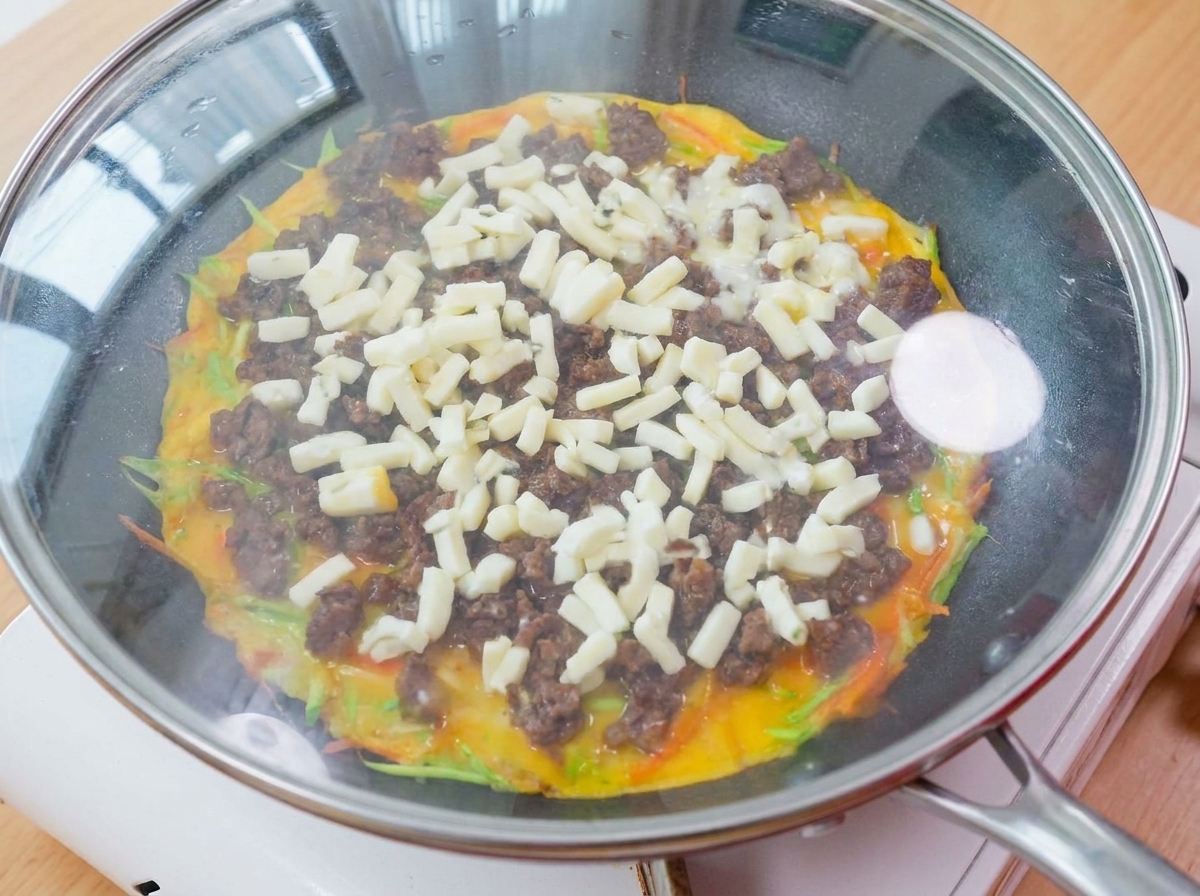 A pan covered with a glass lid resting on a stove, with steam and melting cheese visible inside.